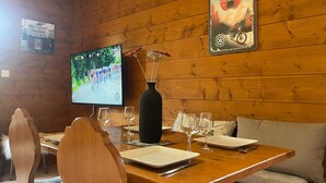 A wooden-paneled room with a dining table set for two, a television displaying a cycling event, and a poster on the wall. - Quiet apartment in Samoëns with pool and WiFi (Samoëns)