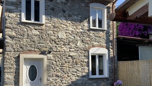 Un edificio in pietra con tre finestre e un portone, un tavolino con un vaso, e un recinto di legno decorato con fiori viola.