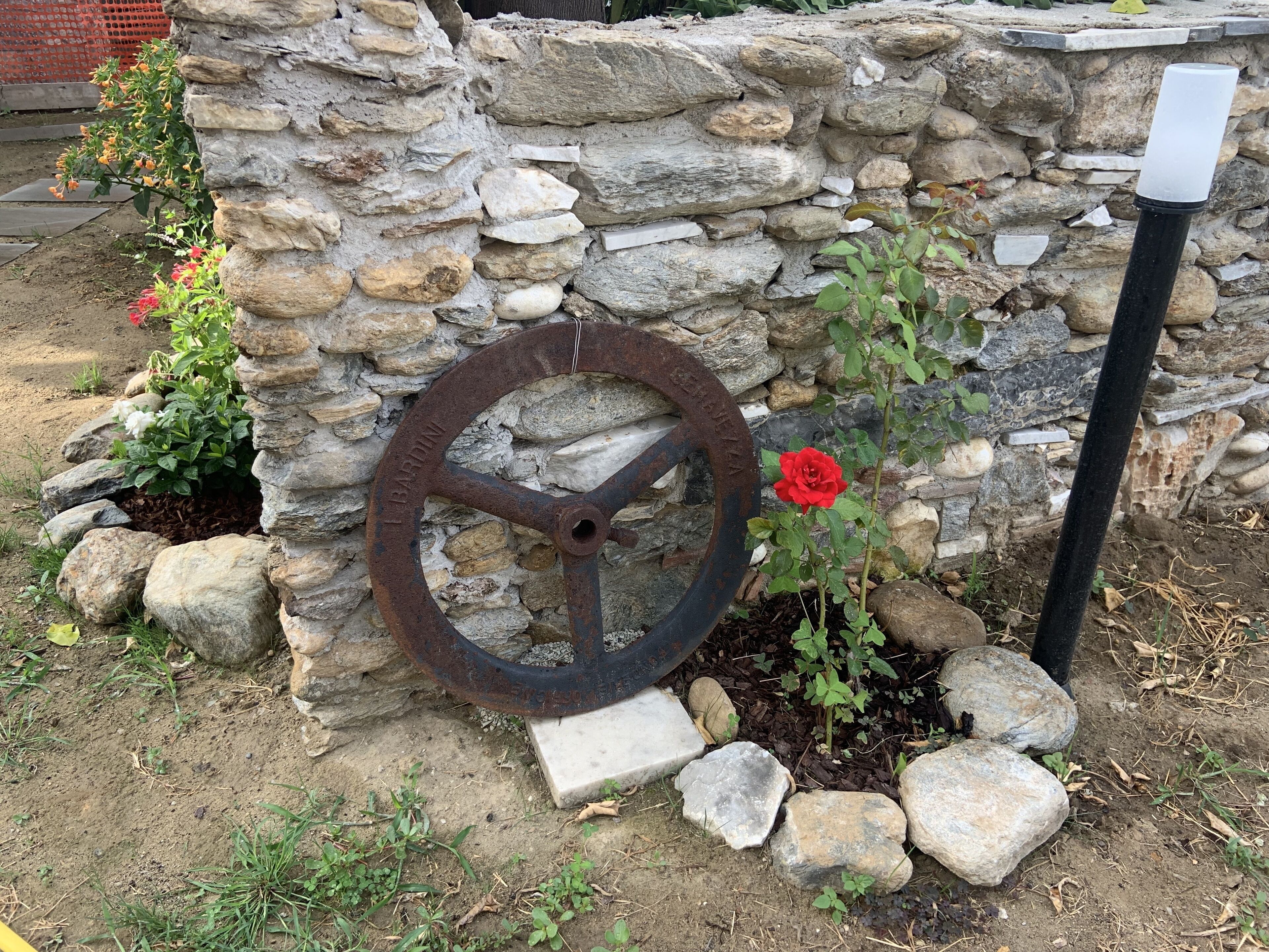 Een ruwe stenen muur met een groot metalen wiel, een rode bloem en een zwarte pijp.