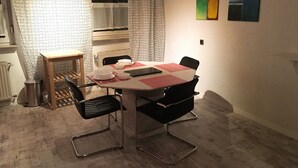 A dining area with a table set for four, surrounded by black chairs with metal frames. A small wooden table with a metal tray is to the left. The room has patterned curtains and a radiator on the wall.
