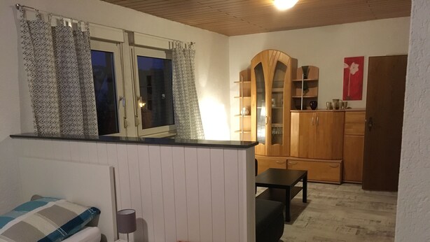 A small, cozy room with a bed, a bedside table with a lamp, a wicker basket, a small table, and a kitchenette area with wooden cabinets and a countertop.