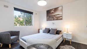A modern bedroom with a bed, two bedside tables, a chair, a large window, and a wall-mounted artwork.