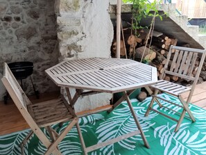 A wooden table and chairs set on a green patterned rug, with a stone wall and a stack of firewood in the background.