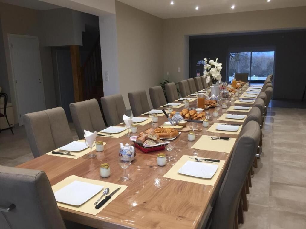 Una larga mesa de comedor dispuesta con platos, cubiertos y bebidas, rodeada de sillas en una habitación moderna con un ventanal grande y una escalera.