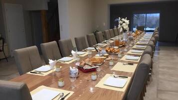 Una larga mesa de comedor dispuesta con platos, cubiertos y bebidas, rodeada de sillas en una habitación moderna con un ventanal grande y una escalera.