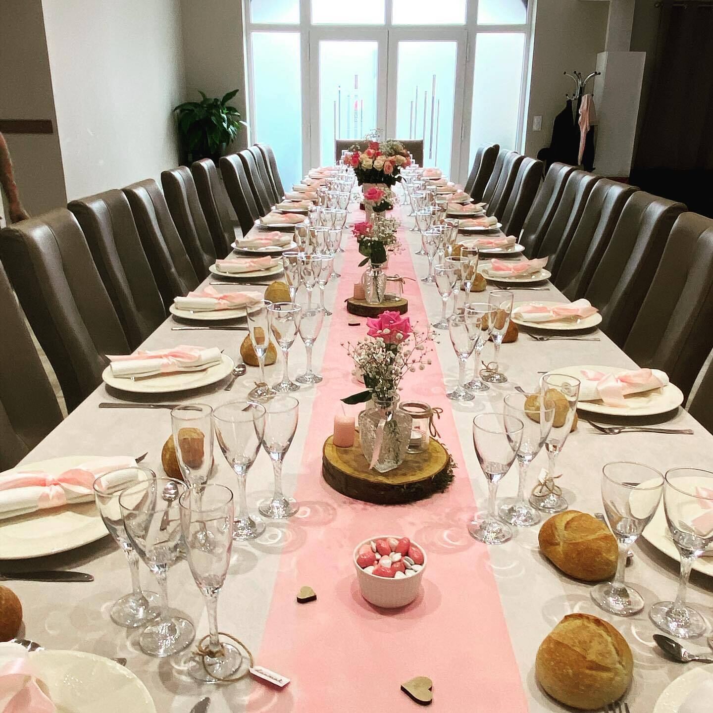 A long dining table set with pink tablecloth, glassware, plates, and bread rolls, flanked by leather chairs.