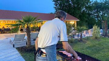 Ein Mann grillt Wurst draußen, im Hintergrund sind ein Haus und eine Terrasse zu sehen.