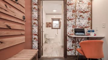 A bathroom with a toilet, a sink, and a shower area. There is a wooden bench and a chair in the room.