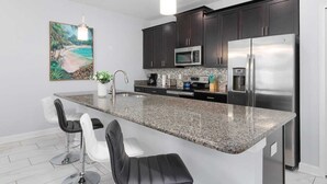 A modern kitchen with dark cabinets, stainless steel appliances, a granite countertop, and bar stools. - Storey Lake Home w/ Pool | Mins to Disney | 4BR | Waterpark (Kissimmee)