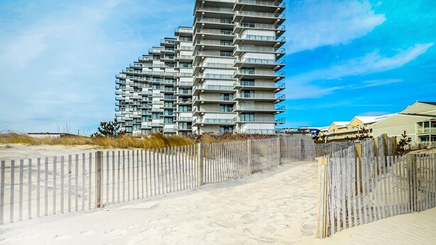 Een modern hoog gebouw met meerdere balkons, gelegen aan een zandstrand met een houten hek op de voorgrond.