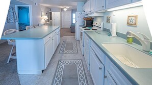 Een keuken met witte kastjes, een spoelbak, een magnetron en een fornuis. Er ligt een vloerkleed en er staat een stoel in de hoek.
