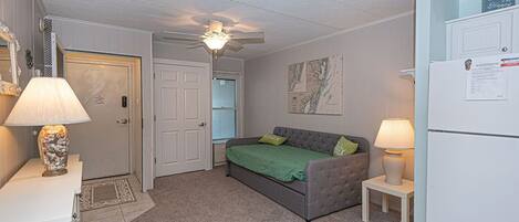A small, well-lit bedroom with a bed, a ceiling fan, a refrigerator, and a door leading to another room.