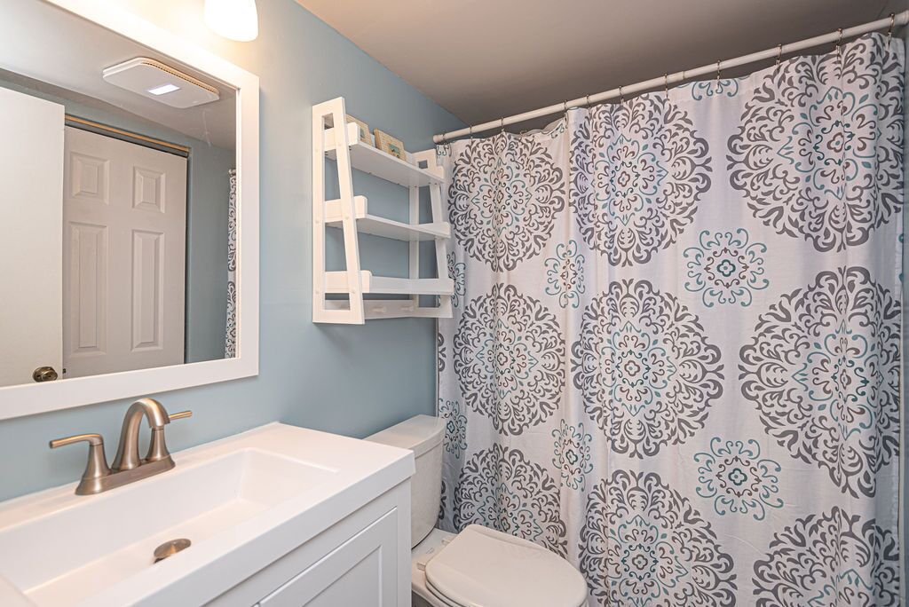 Een badkamer met een wit wastafelmeubel, een wastafel, een spiegel, een toilet en een douchegordijn met een bloemenprint.
