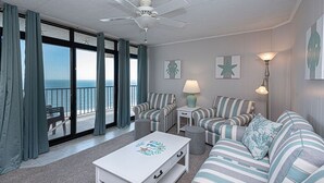 Een woonkamer met gestreepte banken, een witte salontafel en een plafondventilator. Twee lampen en een uitzicht op de oceaan door grote ramen.