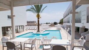 Un espace piscine agrémenté de chaises longues et de tables blanches, d’un palmier et offrant une vue sur le paysage urbain.