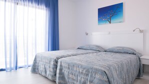 Une chambre avec deux lits, une grande fenêtre avec des rideaux bleus et un tableau représentant un arbre accroché au mur.