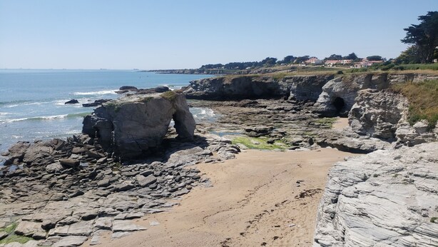 En stenet strand med en naturlig klippeformation, en sti af sand og udsigt til havet og fjerne bygninger.