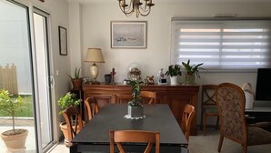 Et spiseområde med et bord, stole og en lysekrone. Der er et vindue med persienner og en dør, der fører ud til en balkon med krukker med planter.