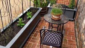 Une petite terrasse extérieure avec un sol en briques, une table et une chaise en métal, et divers pots de plantes.