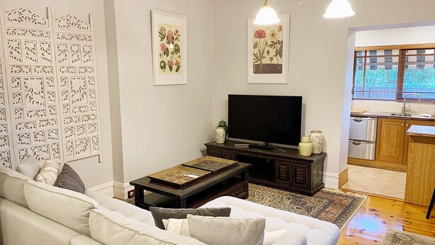 Un salon comprenant un canapé, une table basse, un téléviseur, des œuvres d’art décoratives accrochées au mur, et un coin cuisine visible à travers une porte ouverte.