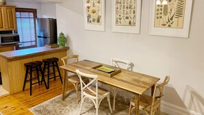 Un coin repas avec une table en bois, des chaises blanches et un tapis. Des reproductions botaniques encadrées ornent le mur.