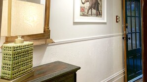 Un couloir avec une armoire en bois, un miroir, un tableau représentant un éléphant encadré, une lampe verte suspendue et une porte bleue.