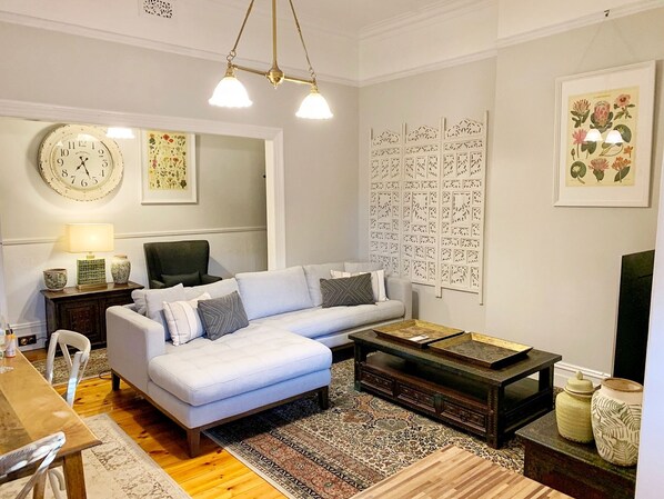 A living room with a light blue sofa, a wooden coffee table, a patterned rug, a wall clock, and a decorative panel with botanical illustrations.