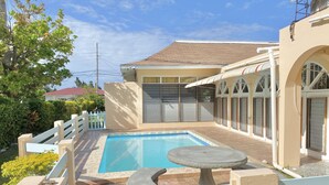 Una piscina con una mesa de piedra, rodeada por una valla blanca y un edificio color beige con ventanas arqueadas.