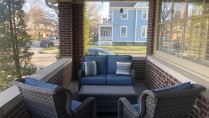 A covered porch with wicker furniture and a view of a blue house in the background. - The Brickmason’s House (Racine)