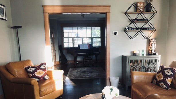 A living room with two brown leather sofas, a round wooden coffee table, and patterned throw pillows. A piano is visible in the background. - The Brickmason’s House (Racine)