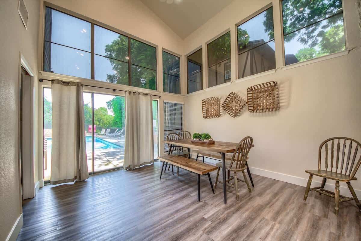 A dining area with a wooden table, chairs, and a bench. Large windows provide natural light and a view of the outdoors.
