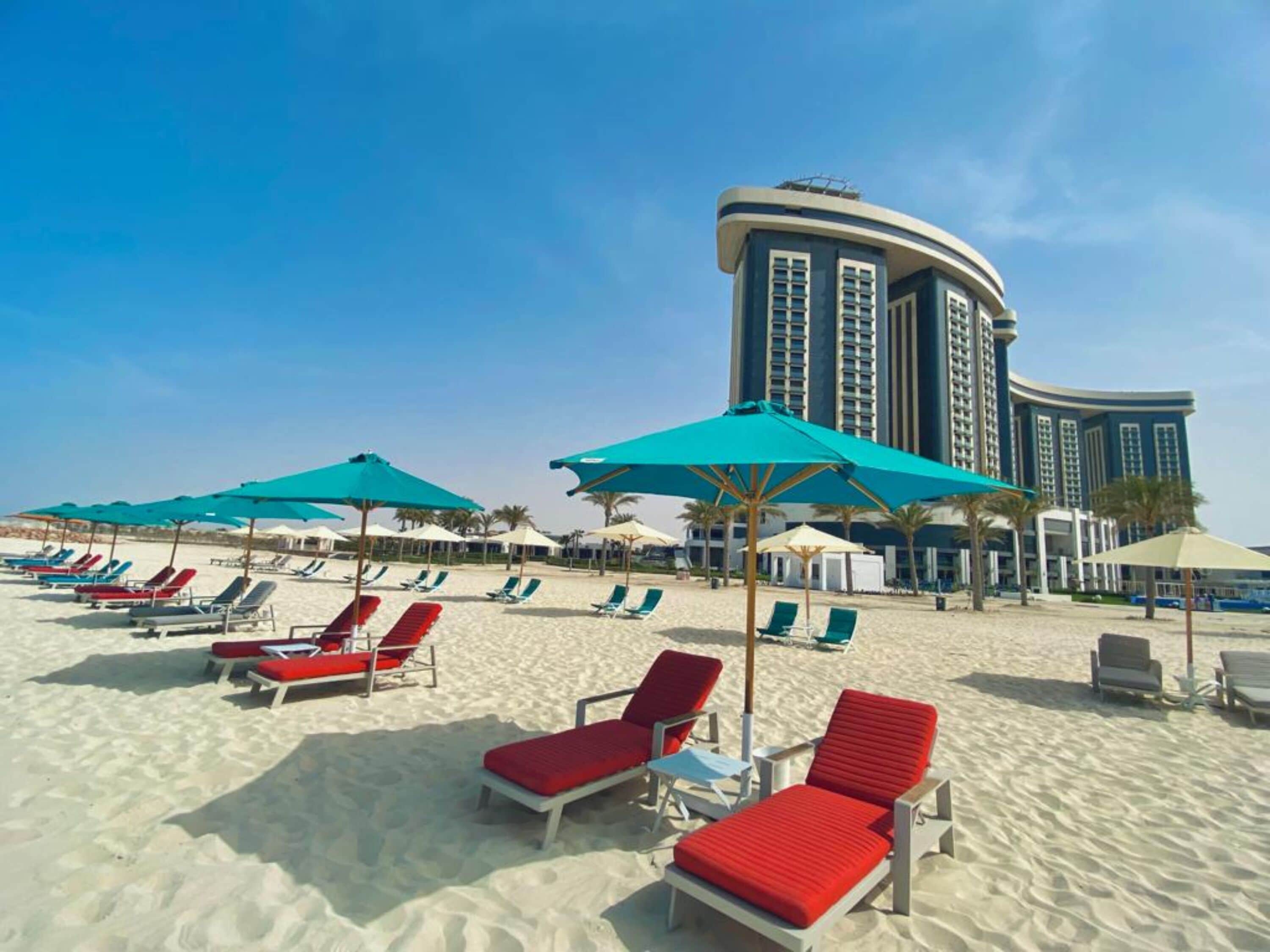 Uma praia com fileiras de espreguiçadeiras e guarda-sóis, com um edifício alto de hotel ao fundo.