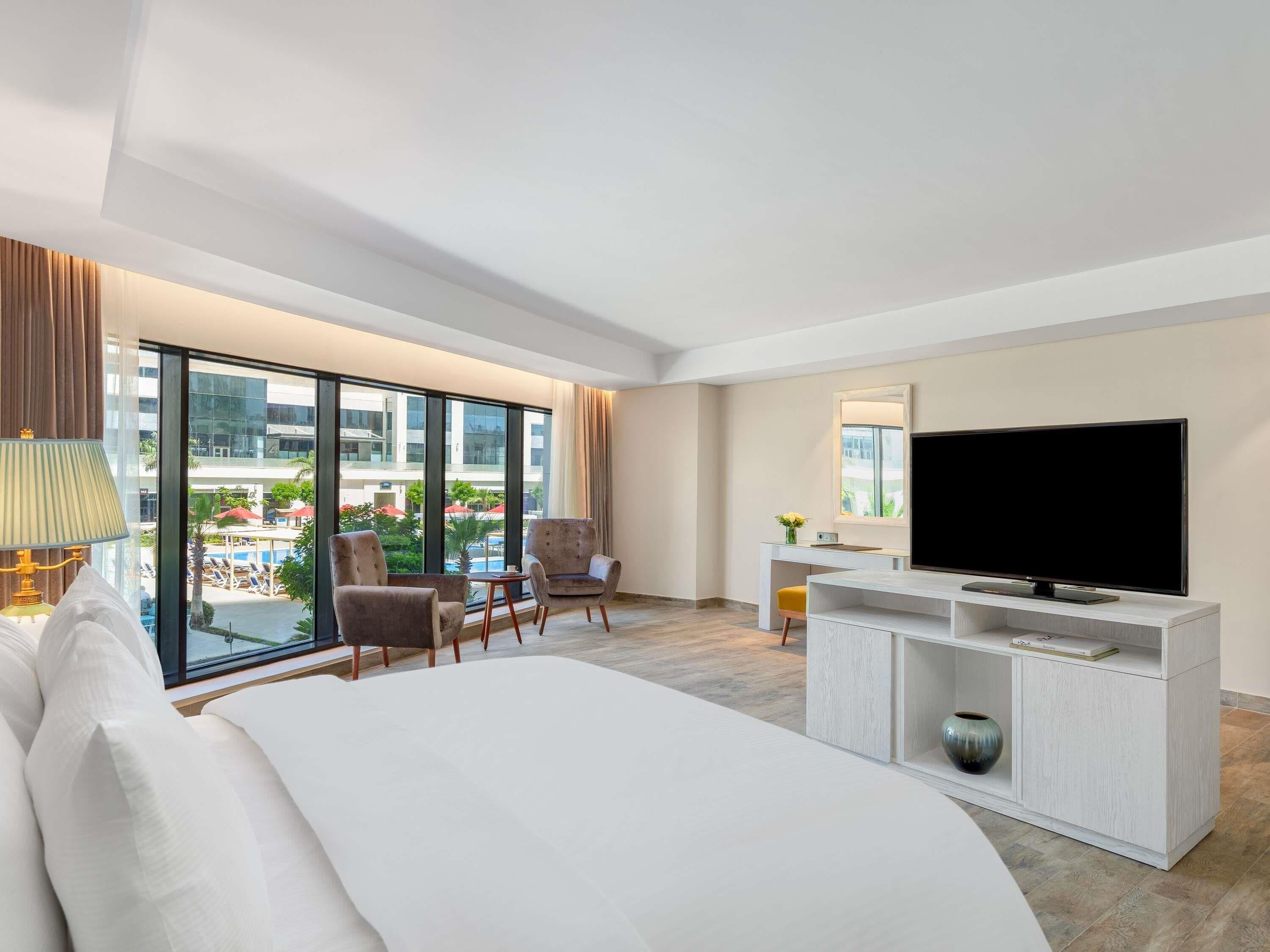 A hotel room with a bed, two armchairs, a TV, a desk, and a large window with a view of a pool and buildings.