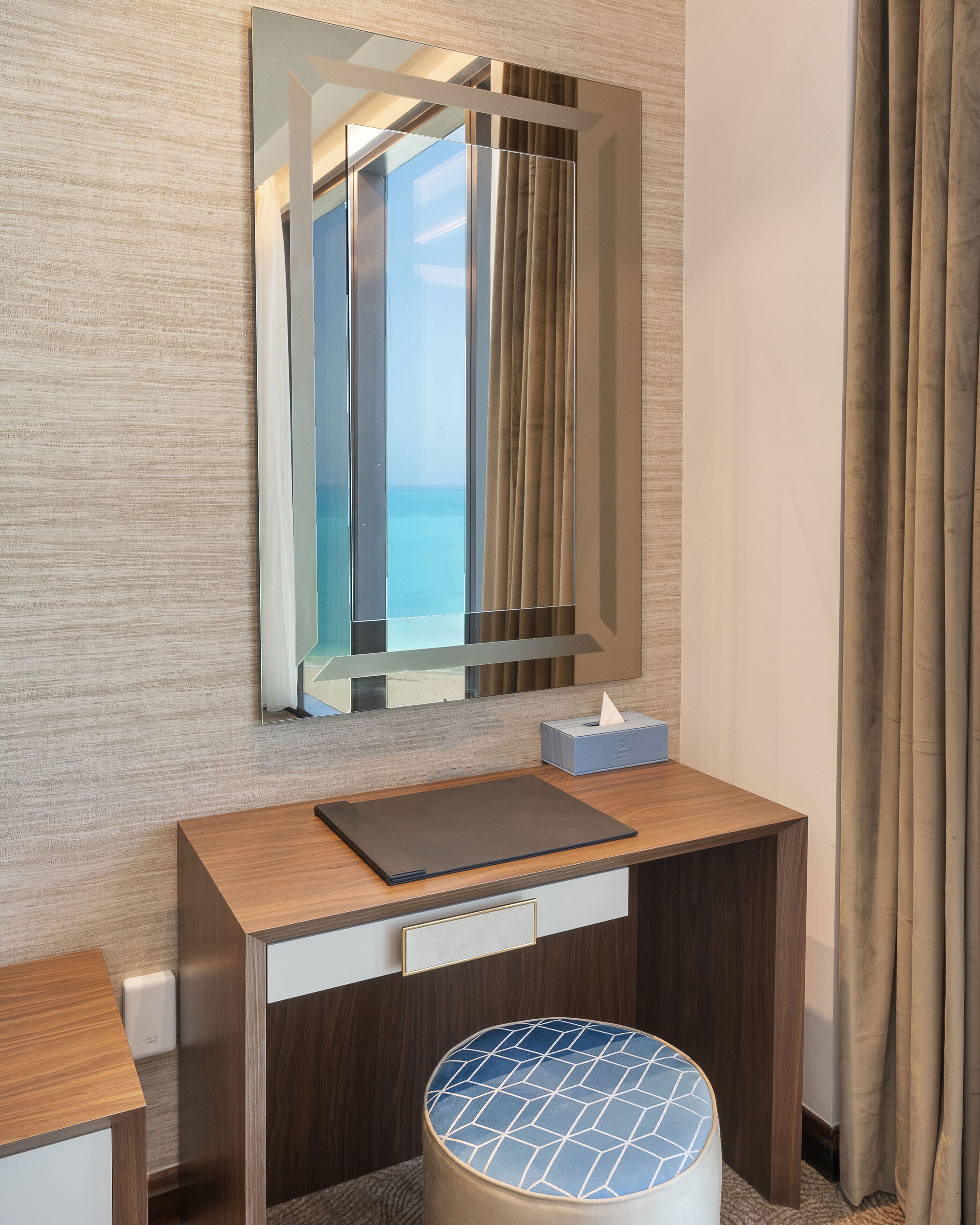Een hotelkamer met een houten bureau, een spiegel, een zakdoosje en uitzicht op zee.