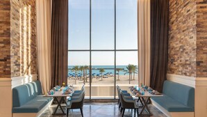 Un área de comedor con vista a la playa, que incluye una mesa larga con sillas, un sofá azul y un amplio ventanal con cortinas.
