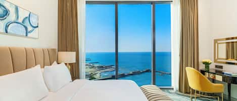 A hotel room with a large bed, a desk with a chair, a mirror, and a view of the sea.
