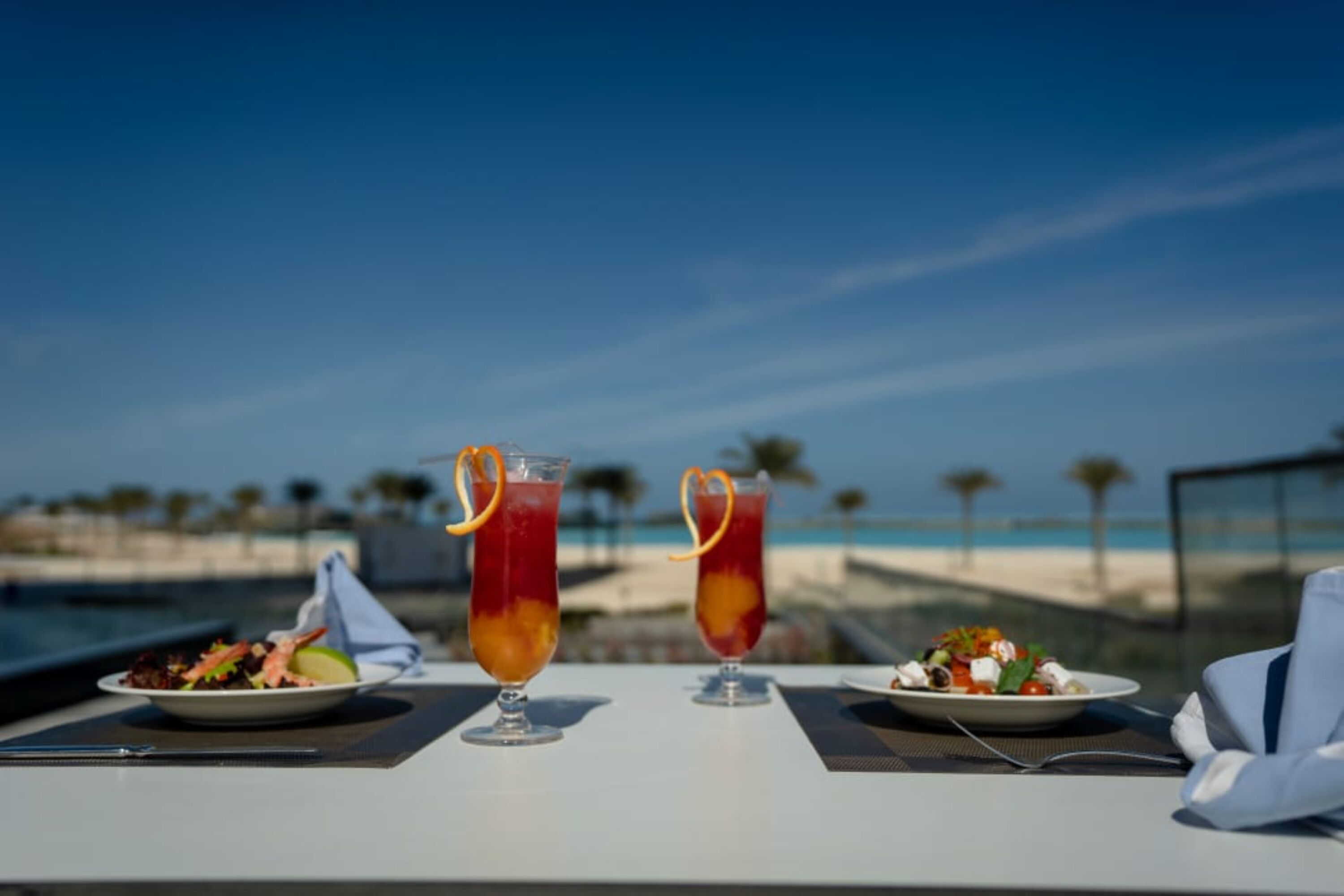 Ein Esstisch mit zwei hohen Gläsern rotem Getränk, einem Salat und einer Speisenplatte, vor einer Strandkulisse.