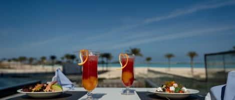 Una mesa de comedor con dos vasos altos de bebida roja, un plato de ensalada y un plato de comida, con vista a la playa.