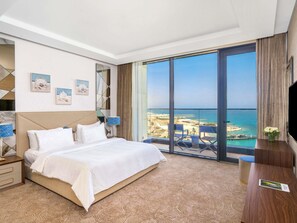 A hotel room with a large bed, a TV, a desk, and a balcony with a view of the sea. - Rixos Premium Alamein (El Alamein)