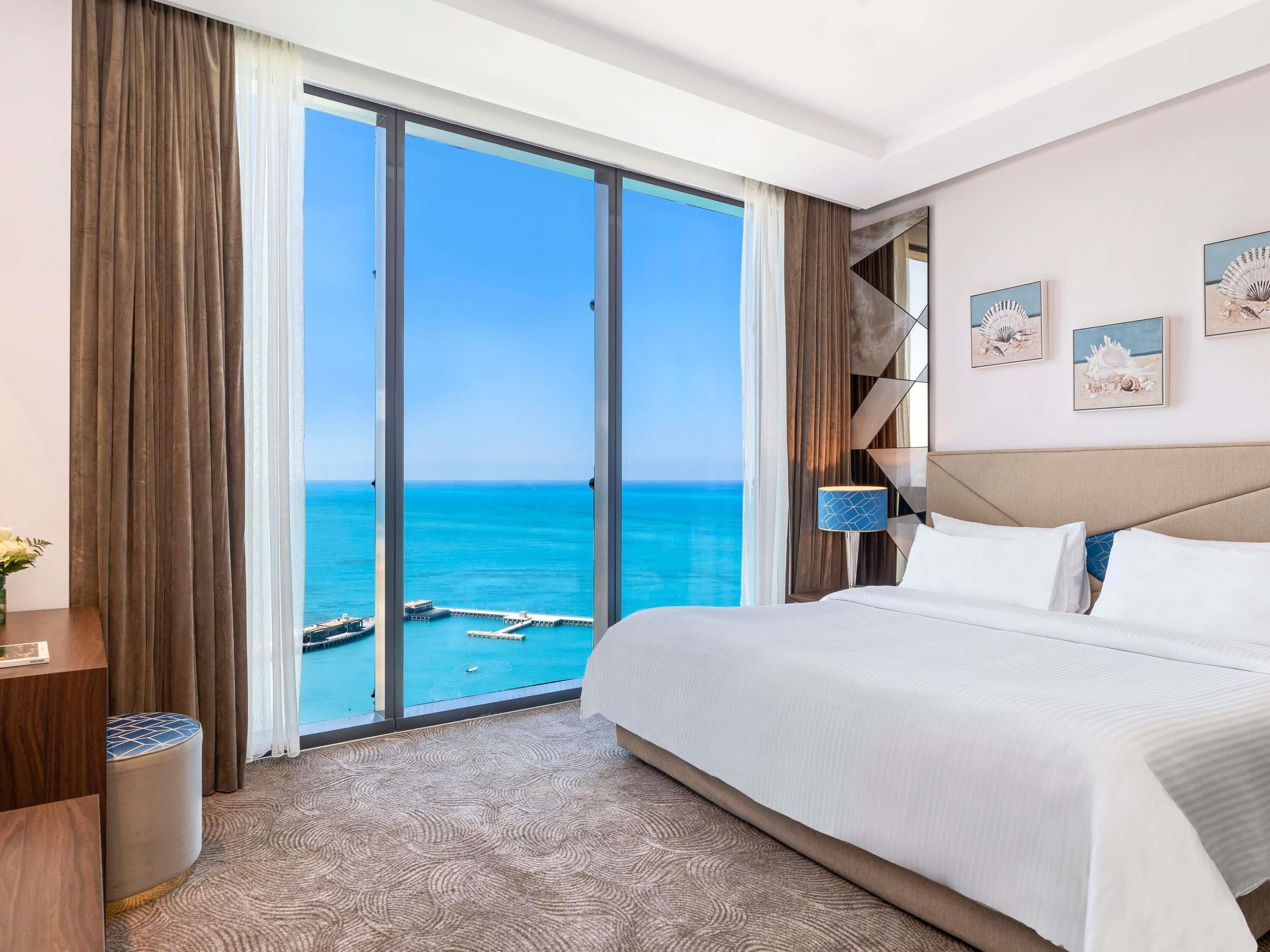 A hotel room with a large bed, a view of the ocean, a wooden side table, and a lamp.