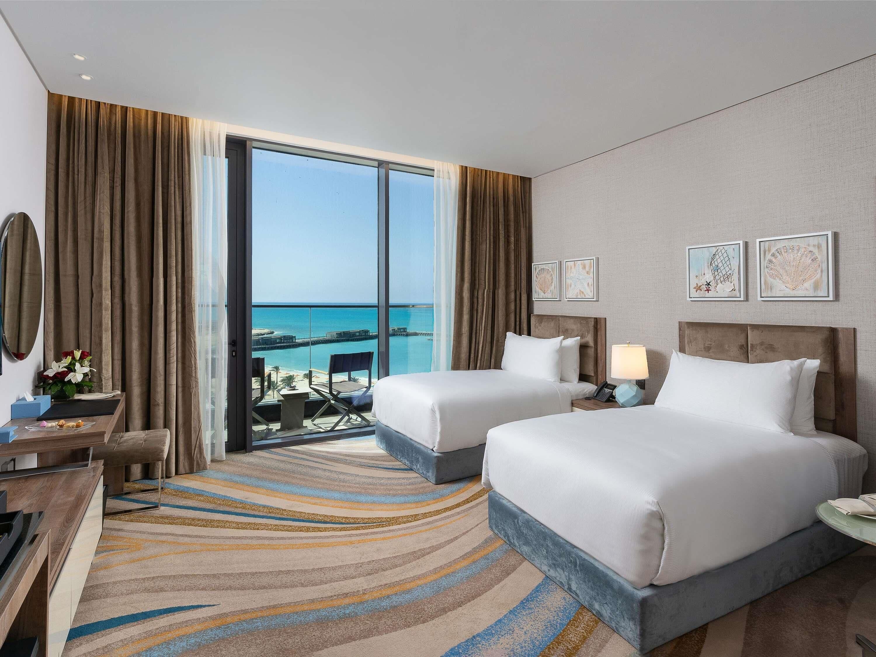 Habitación de hotel con dos camas, un ventanal con vista al mar, un escritorio con una silla y un balcón pequeño.