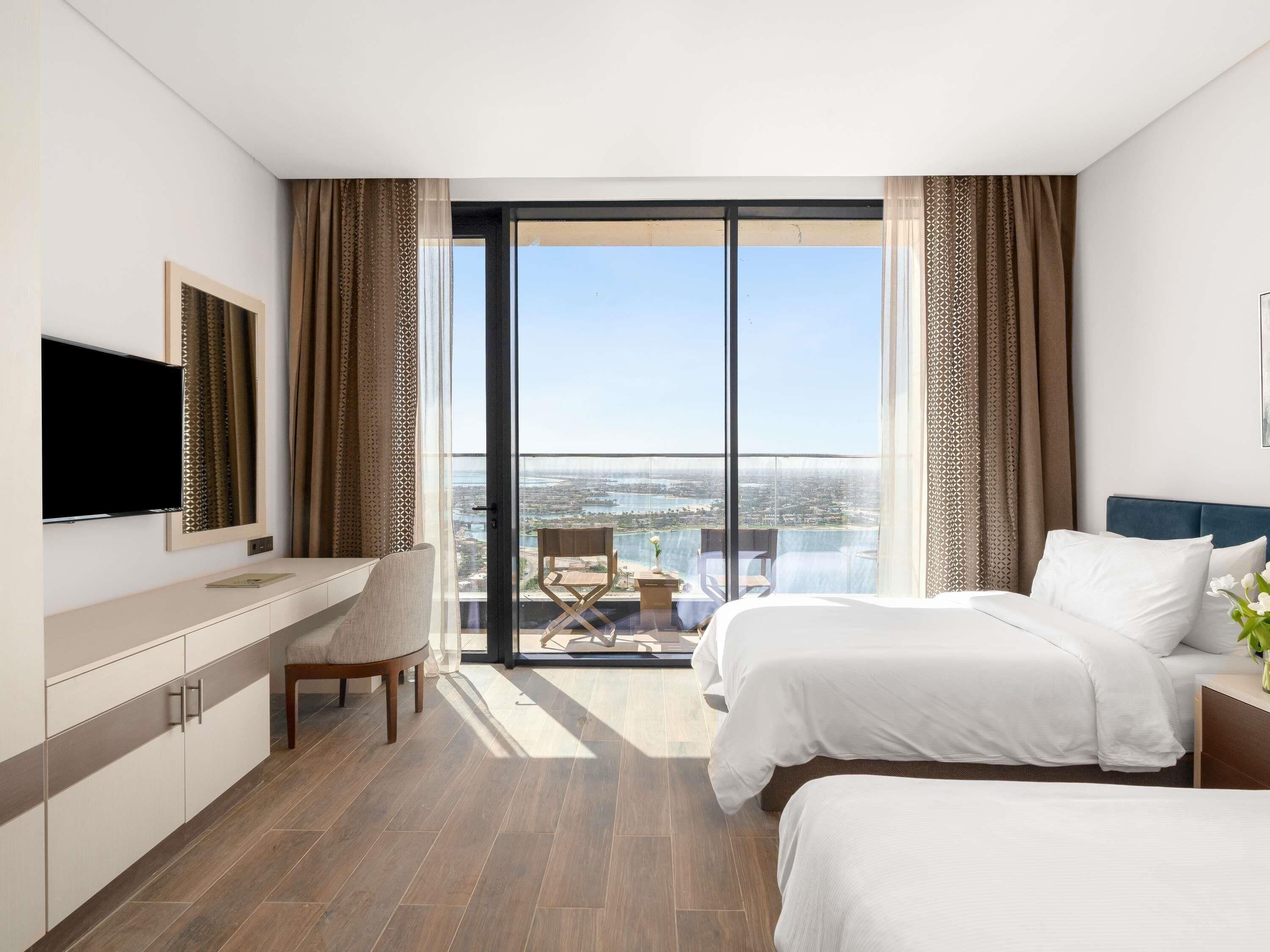 A hotel room with a bed, a desk, a chair, a TV, and a balcony with a city view.