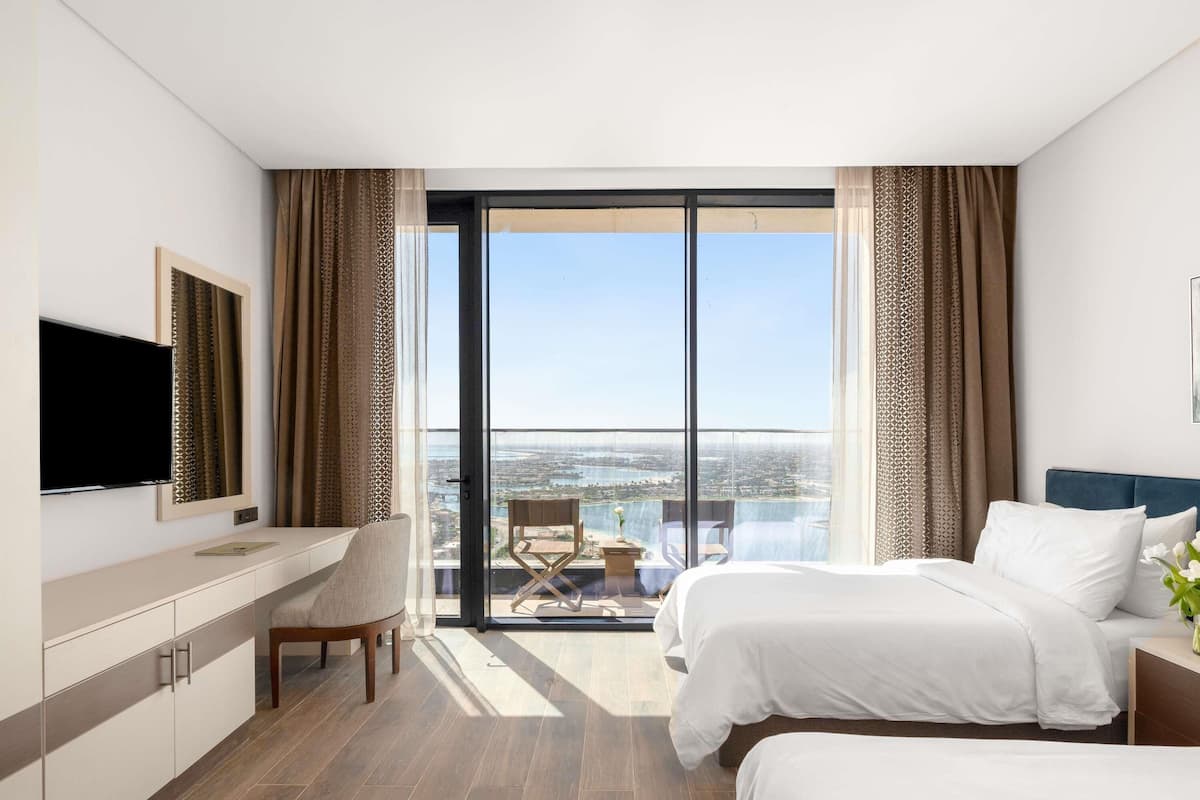 A hotel room with a bed, a desk, a chair, a TV, and a balcony with a city view.