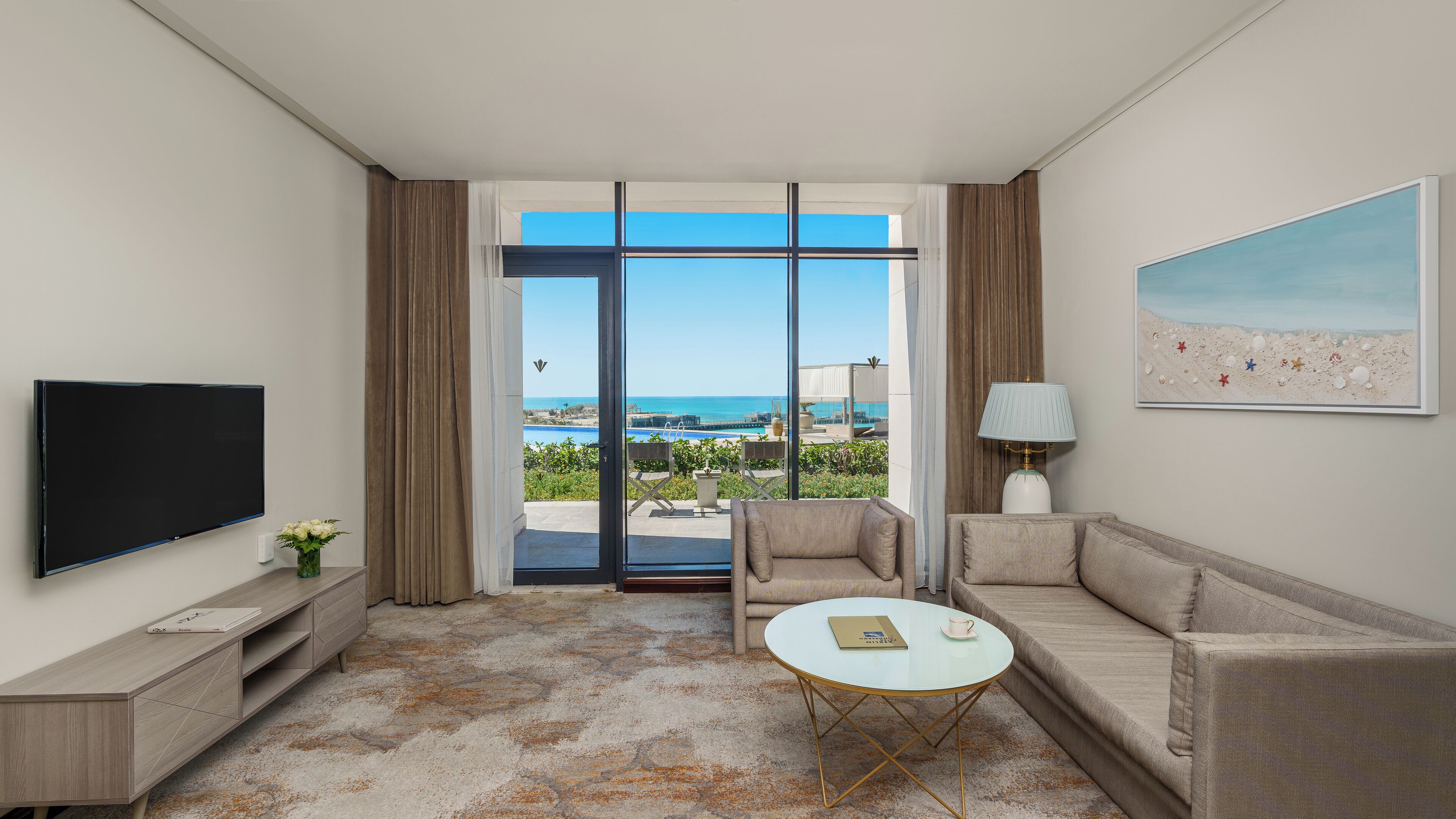 Habitación de hotel con televisor grande, sofá, una mesita y vista a la playa.