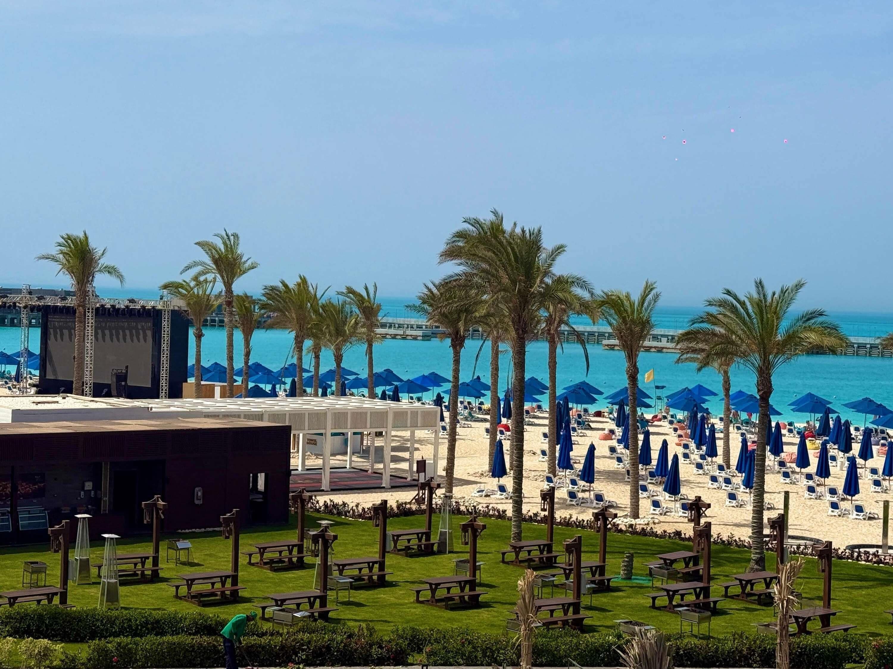 Un hotel frente a la playa con un escenario, palmeras y sillas para tomar el sol.