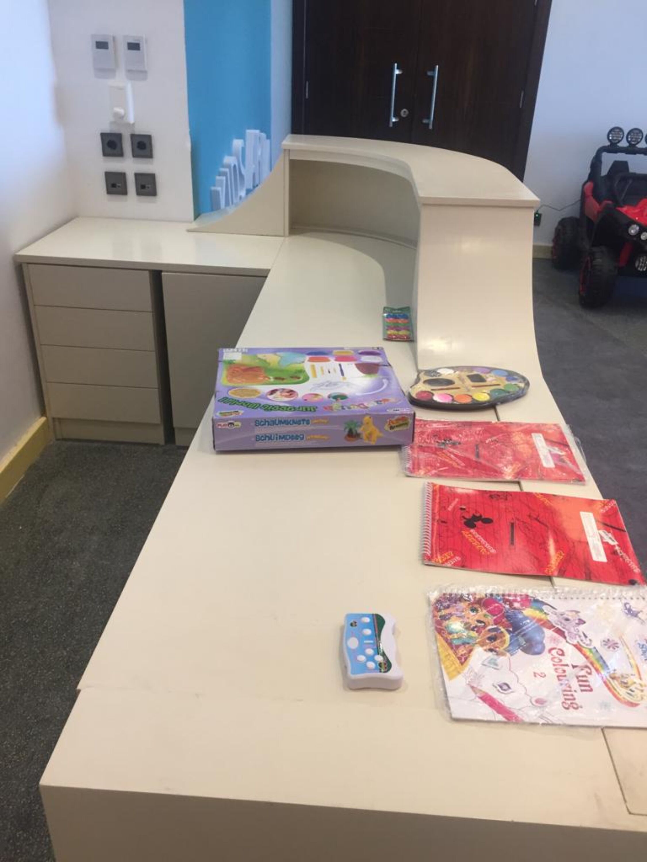 A desk with a box of Schaumkiste coloring books, a remote control, and several coloring books.