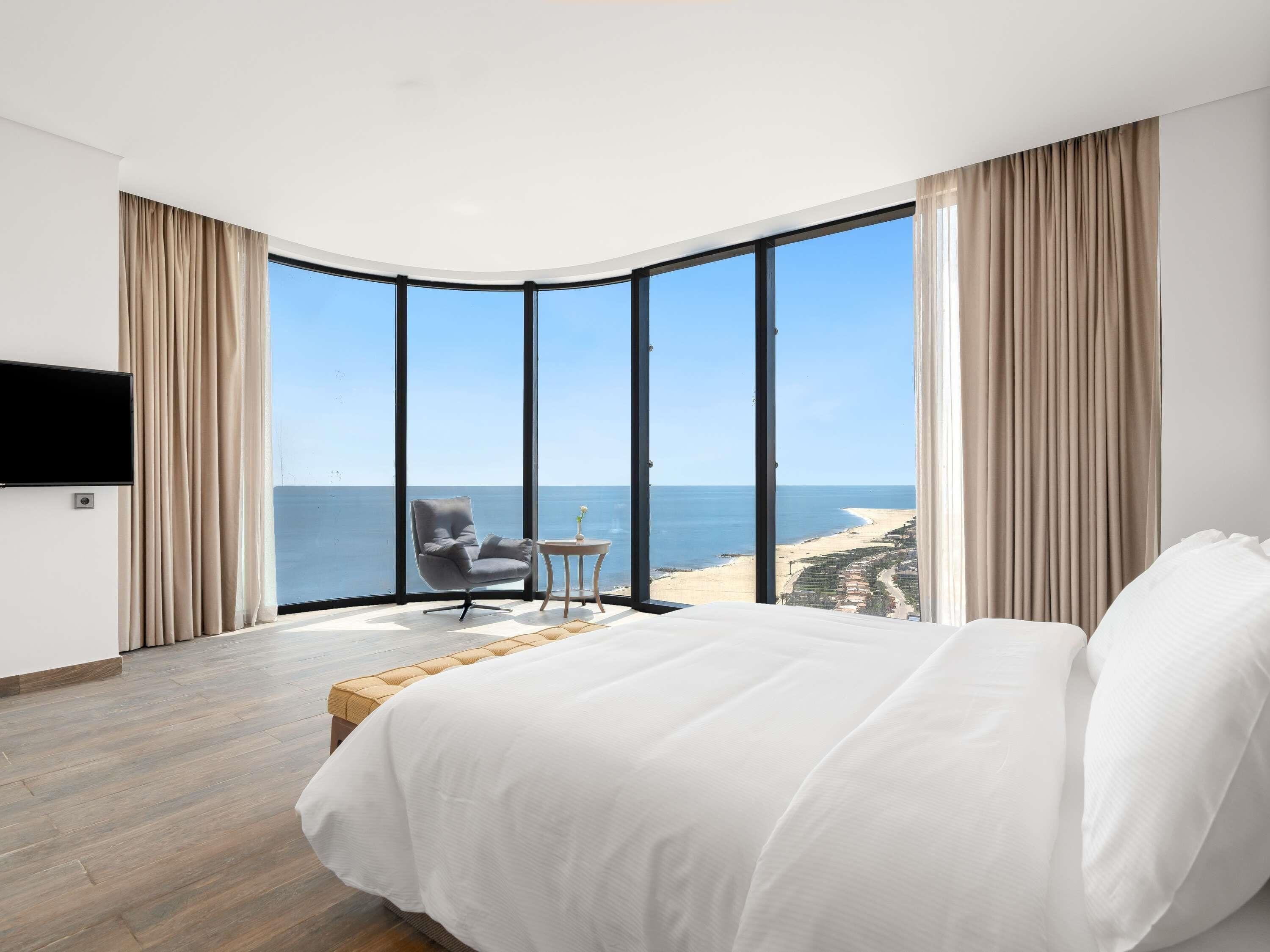 A bedroom with a large bed, a chair, a small table, and a TV. It has a view of the beach and ocean.