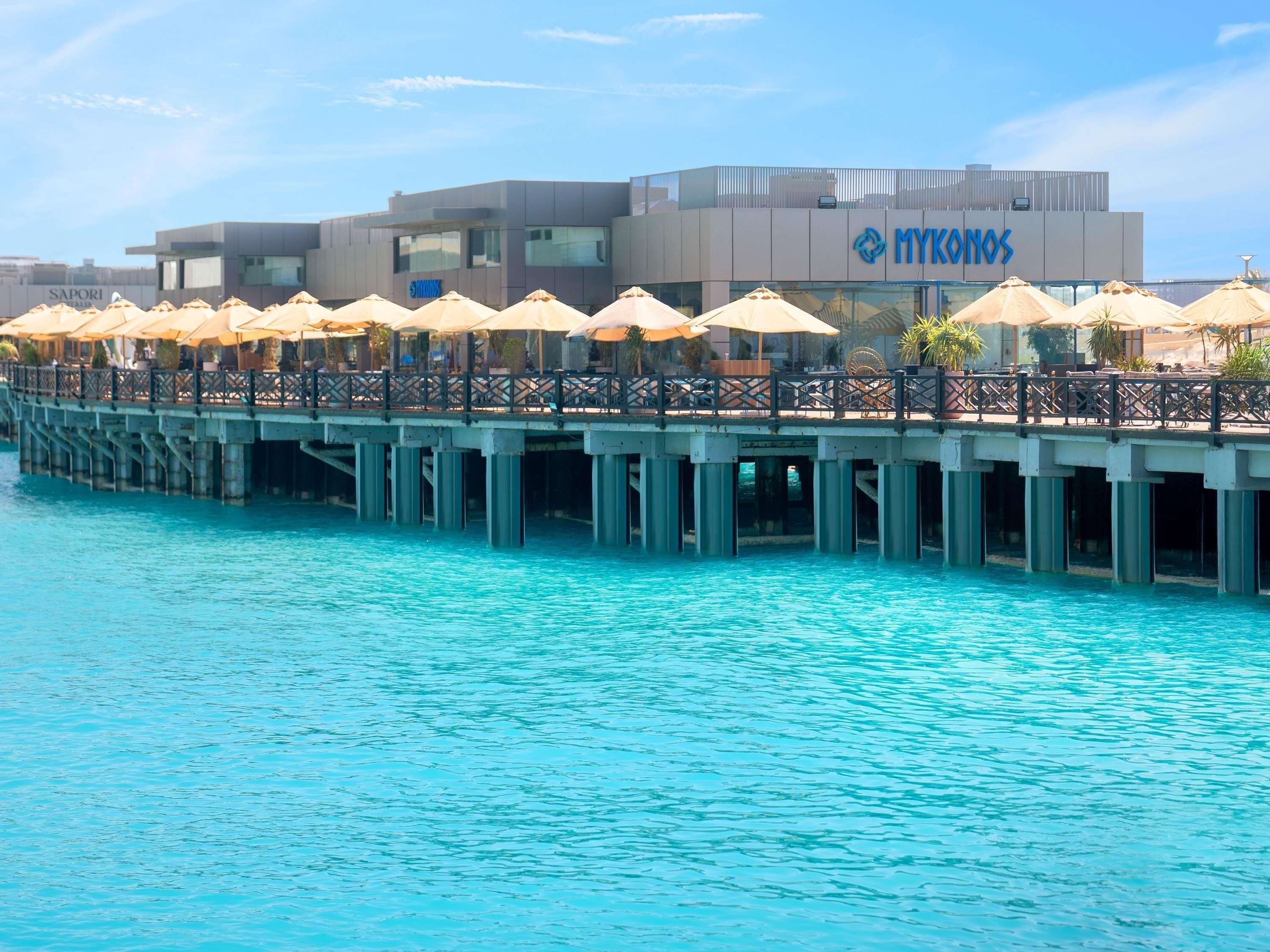 A waterfront hotel with a pier extending over the water, featuring outdoor seating with umbrellas.