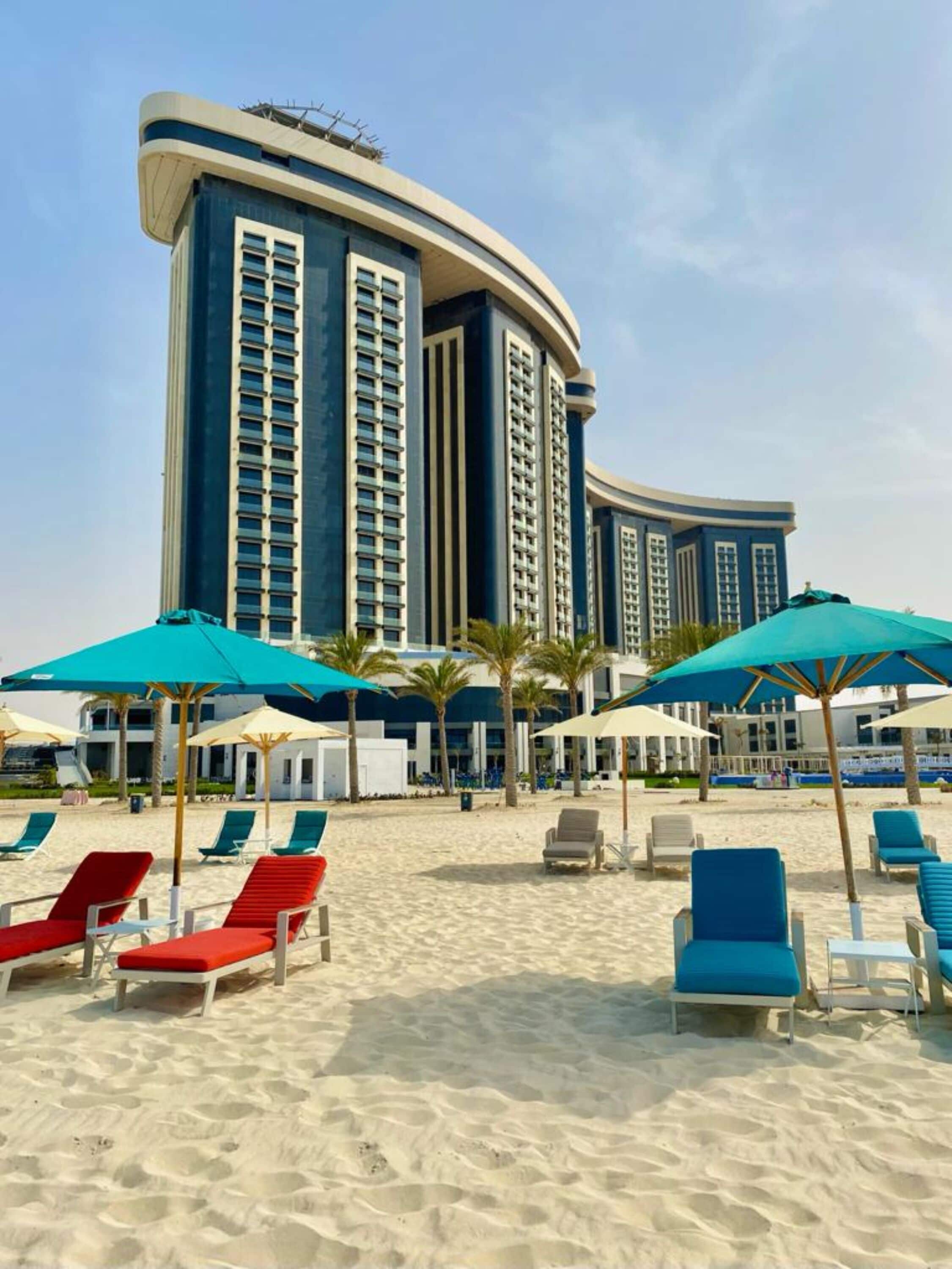 A beachfront hotel with a curved facade, multiple balconies, and a sandy beach area with lounge chairs and umbrellas.