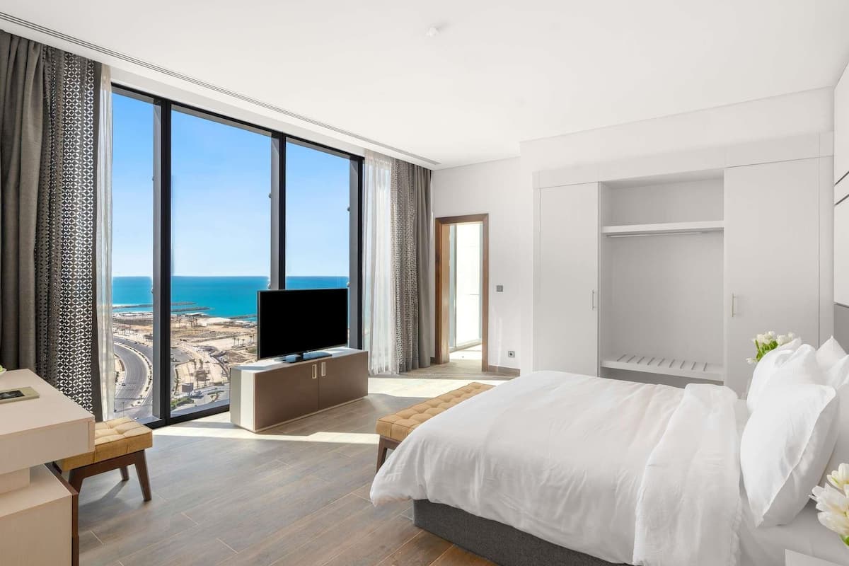 A modern hotel room with a large window offering a view of the coastline, a bed, a TV, and a walk-in closet.